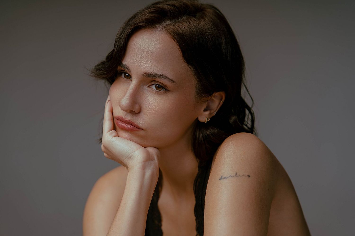 Professional studio portrait in muted tones featuring a woman with dark hair in a contemplative pose, hand resting on chin. The subject wears minimal makeup and a sleeveless top, photographed against a neutral gray background. Soft, professional lighting 