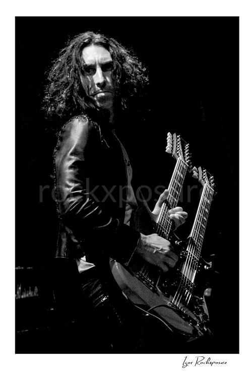 Vertical black and white image of Brent Woods holding a double-neck guitar on stage in a dramatic close-up