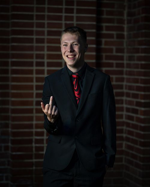 PACI grad giving the finger standing in front of a brick wall in his suit.