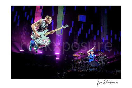 Horizontal color image of Flea and Chad Smith of Red Hot Chili Peppers performing live on stage, with Flea soaring high with his bass while Chad Smith plays drums beneath vivid purple concert light