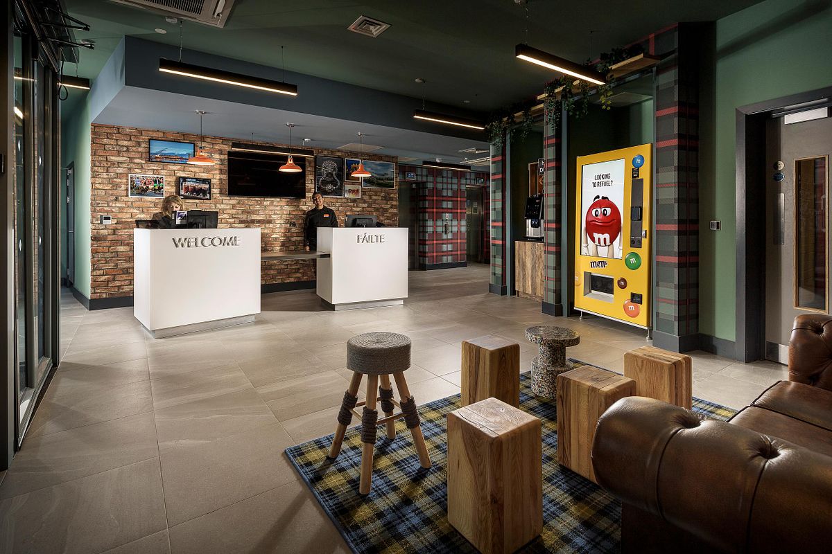 Reception area of the Easy Hotel Dublin. Two white check-in desks with 'WELCOME' and 'FAITE' signs are visible. A large M&M's vending machine is against a green wall with a geometric pattern. Wooden stools and a leather sofa are in the foreground.