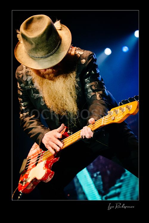 Vertical color image of Dusty Hill of ZZ Top performing live on stage with a red bass guitar, head lowered beneath his wide-brim hat with blue stage lights glowing behind him