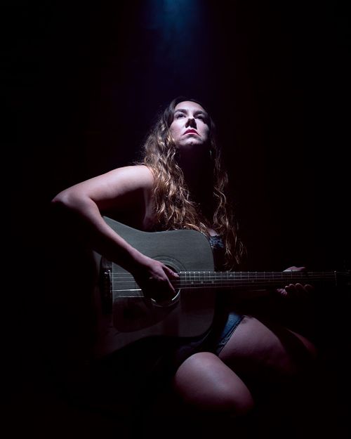 A low key portrait of a woman playing a gray guitar while looking up the light above her.