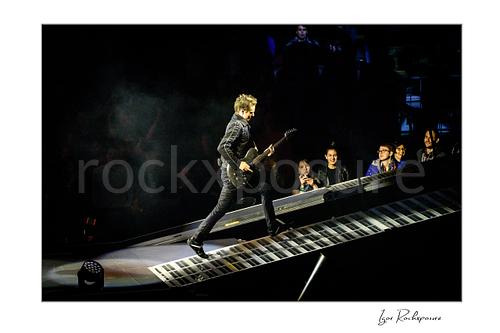 Horizontal color image of Matt Bellamy of Muse walking up a piano-key stage ramp with guitar under dramatic concert lighting
