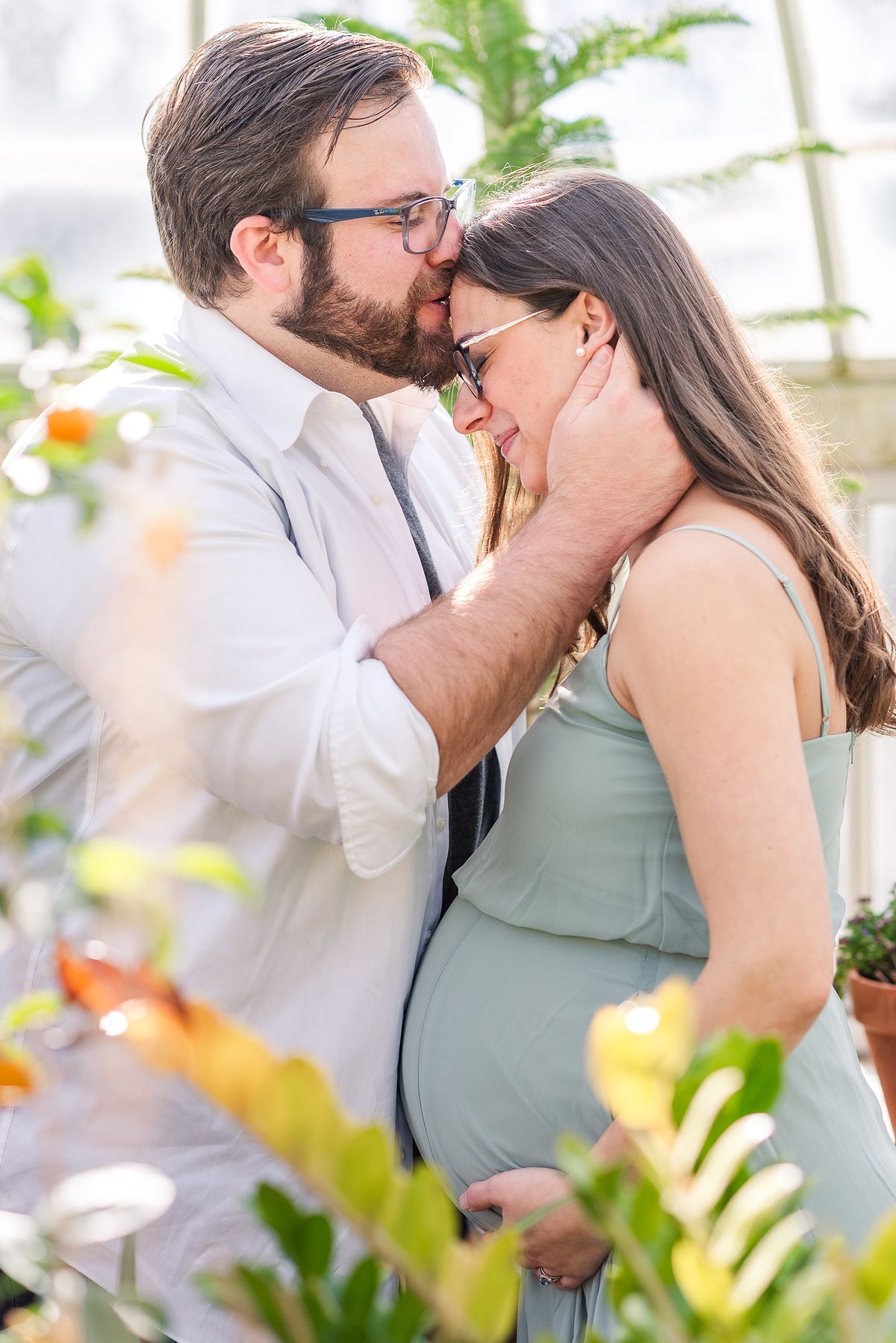 Husband kissing his expectant wife's forehead while she holds her pregnant belly in The Frick greenhouse