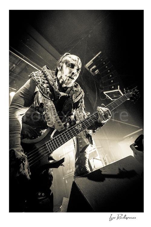Vertical black and white image of Orion of Behemoth playing bass in a dramatic low-angle live performance shot with corpse paint and stagewear