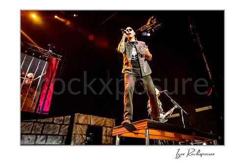 Horizontal color image of M. Shadows of Avenged Sevenfold singing on a stage riser with a microphone under warm concert lighting