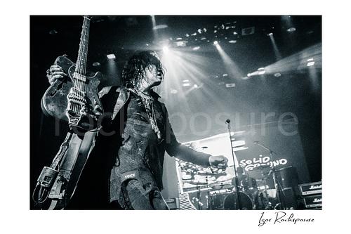 Horizontal black and white image of Tom Keifer on stage holding his guitar upright in a dramatic live performance moment, with bright concert beams and the drum kit visible behind him
