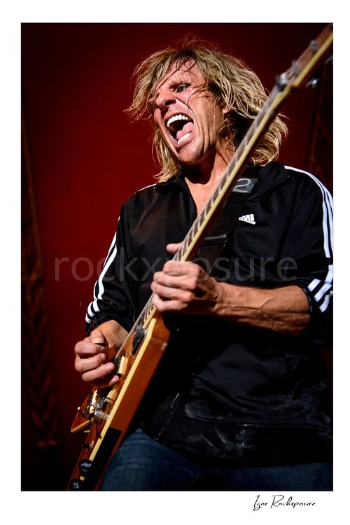 Vertical color image of C.C. DeVille performing with electric guitar during a Poison concert, with wild expression under vivid red stage light