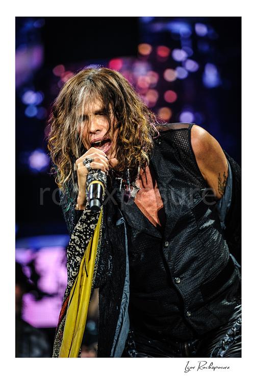 Vertical color image of Steven Tyler of Aerosmith performing live on stage with a handheld microphone, scarf-wrapped mic stand, and tousled hair against colorful blurred lights
