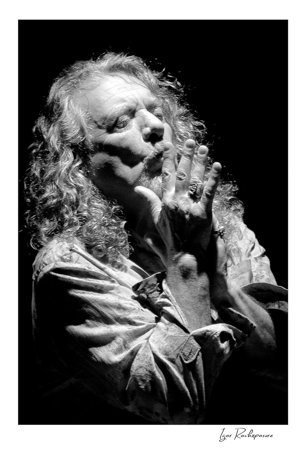 Vertical black and white concert photography of Robert Plant with hands raised near his face in an expressive dramatic portrait under stage lighting