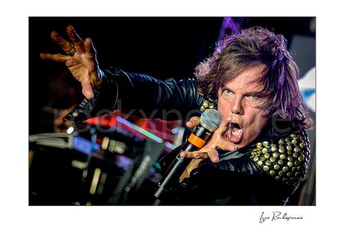 Horizontal color image of Joey Tempest of Europe performing live on stage with a handheld microphone, black leather jacket with studded shoulders, intense expression, and outstretched hand