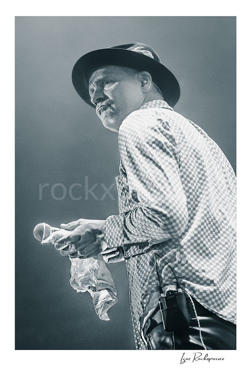 Vertical black and white image of Gord Downie of The Tragically Hip holding a microphone and white cloth in a quiet live performance moment, wearing a hat and patterned shirt under soft stage light