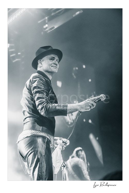 Vertical black and white image of Gord Downie of The Tragically Hip holding a microphone on stage, wearing a hat and leather jacket with bright concert haze and a blurred band member behind him