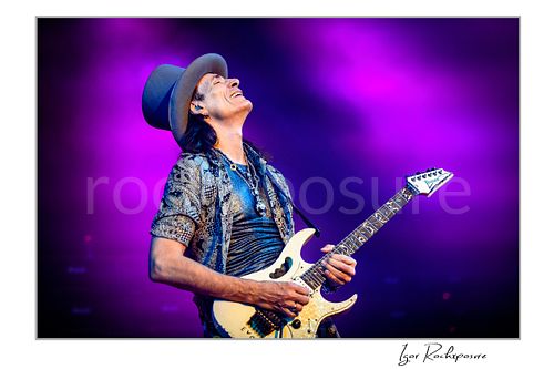 Horizontal color image of Steve Vai playing guitar with head tilted back under vivid purple stage lighting
