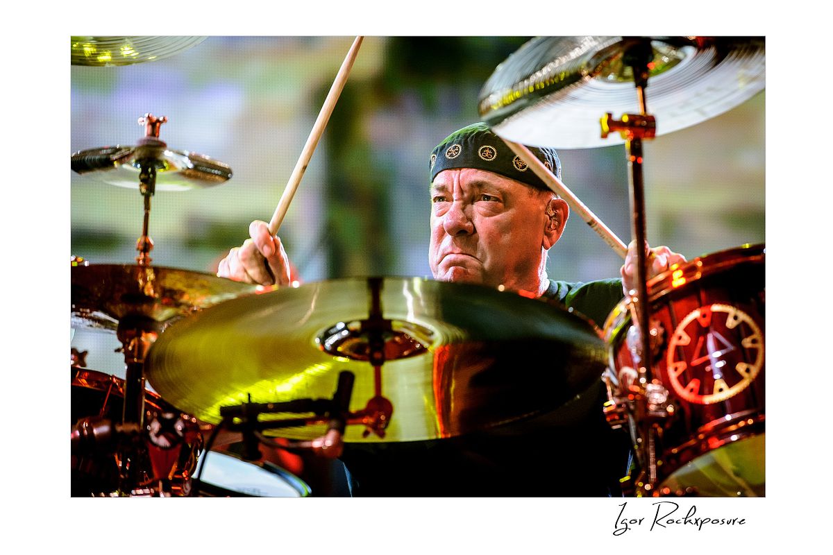 Horizontal concert photography of Neil Peart striking the drums with raised drumsticks surrounded by a large drum kit under warm stage lighting