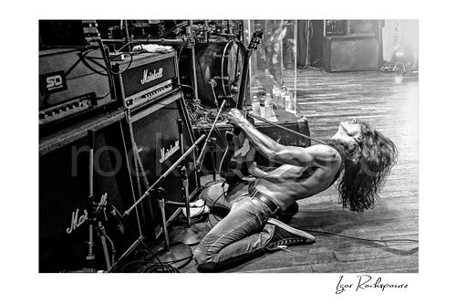 Horizontal black and white image of Brent Woods playing guitar shirtless on the stage floor beside Marshall amplifier stacks