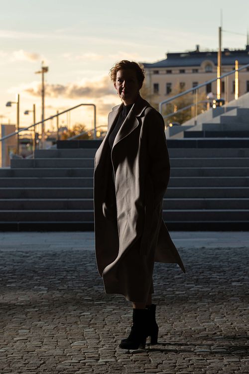 Helkroppsporträtt i motljus på trappa i solnedgång, kvinna i lång kappa med tydligt ljus och skugga, Stockholm.