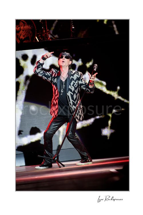 Vertical color image of Klaus Meine of Scorpions performing live in patterned jacket with microphone stand and bold black-and-white stage screen