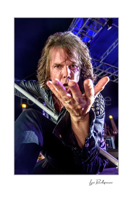 Vertical color image of Joey Tempest of Europe performing live on stage in a black leather jacket with a white microphone stand, dramatic low-angle view, and outstretched hand reaching toward the camera