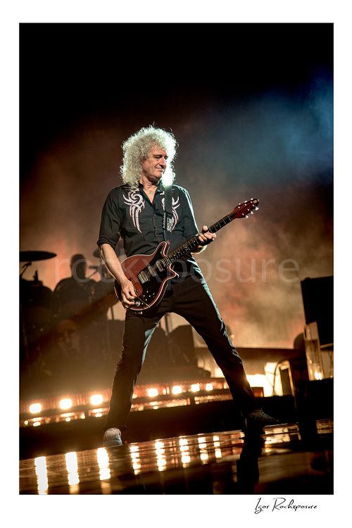 Vertical color image of Brian May of Queen playing guitar onstage in a wide stance with glowing stage haze and warm floor lights