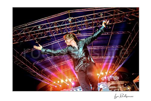 Horizontal color image of Joey Tempest of Europe performing live on stage with outstretched arms, black leather shirt, and bright concert lighting beneath a large outdoor truss roof