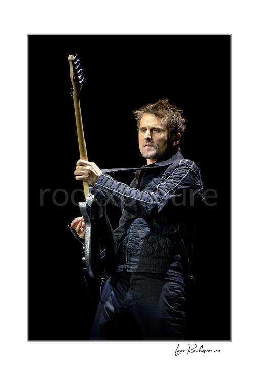 Vertical color image of Matt Bellamy of Muse holding guitar upright in a dramatic stage portrait against a dark background