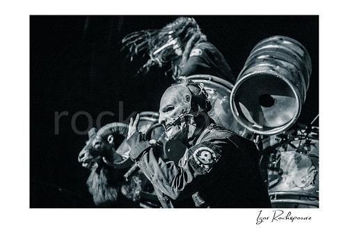 Horizontal black and white image of Corey Taylor of Slipknot wearing a mask and singing into a handheld microphone with industrial drums and a ram prop behind him