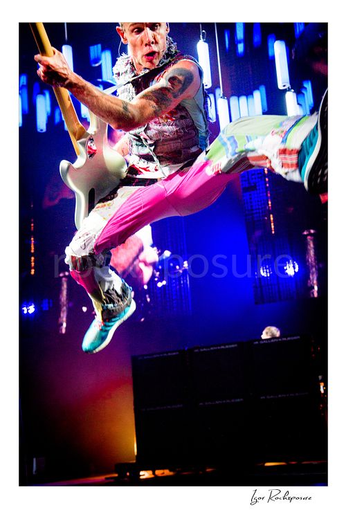 Vertical color image of Flea of Red Hot Chili Peppers suspended midair in a dramatic live performance leap, playing bass with one leg extended against a vivid LED stage backdrop