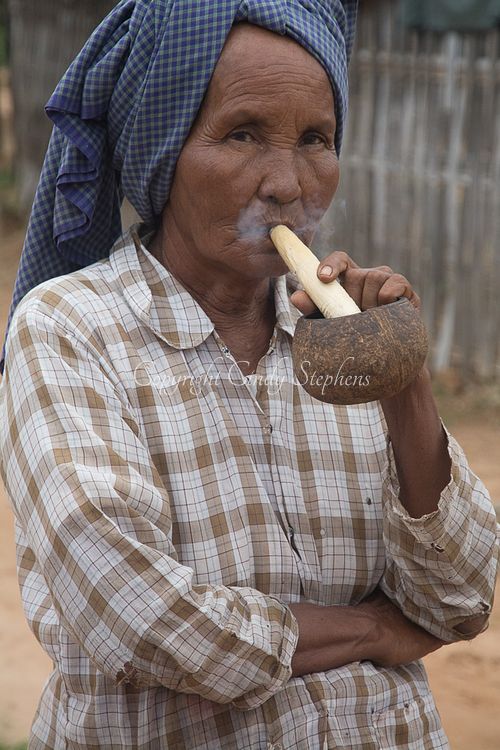 Burmese woman smoking a traditional corn husk pipe while wearing a plaid shirt and turban, with a wooden fence and dirt road in the background