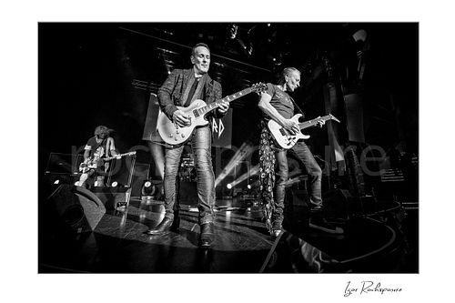 Horizontal black and white image of Rick Savage, Vivian Campbell, and Phil Collen of Def Leppard performing live on stage, with Savage at left on bass, Campbell at center on guitar, and Collen at right on guitar